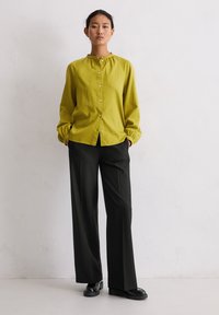 Vrouw die tegen een witte muur staat, gekleed in een felgroen knoopblouse, zwarte wijde broek en zwarte schoenen, met handen in haar zakken.