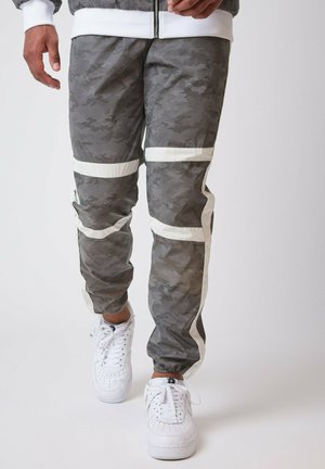 Pantaloni jogging grigi in camouflage con strisce bianche ai lati, realizzati in tessuto leggero. Presentano un cinturino elastico in vita e polsini a gamba affusolata.