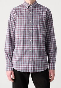 Mann trägt ein aufgeknöpftes, langärmeliges, rot-blau kariertes Hemd mit einer Tasche auf der linken Brust und schwarze Hose.