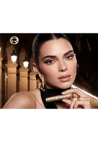Zwarte zijden sjaal om de nek gewikkeld; houdt een gouden, cilindervormige mascara-tube vast met het label "PANORAMA." Gouden oorbellen accentueren de look.