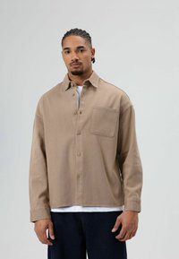 Homme aux cheveux tressés portant une chemise beige boutonnée par-dessus un haut blanc et un pantalon foncé, debout devant un fond clair uni.
