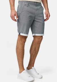 Shorts pour hommes bleus à motifs avec revers roulés, fabriqués en tissu léger, avec poches avant et fermeture à boutons. Portés avec des baskets blanches.