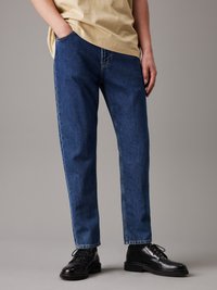 Blå denimjeans med rak passform, fem fickor och minimala sömdetaljer. Bärs med svarta snörskor och en ljusbeige skjorta.