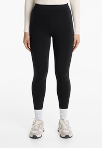 Fekete leggings magas derékkal, sima textúrával, hosszú fazonban, világos színű sportcipővel és fehér bokazoknival párosítva.