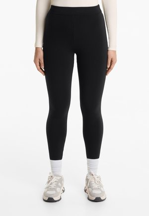 Zwarte leggings met een hoge tailleband, gladde textuur, volledig lengte ontwerp, gecombineerd met lichtgekleurde sportschoenen en witte enkelsokken.
