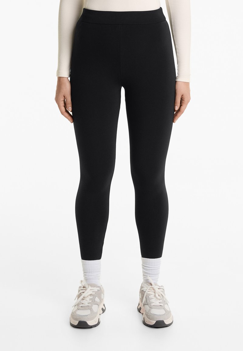 Fekete leggings magas derékkal, sima textúrával, hosszú fazonban, világos színű sportcipővel és fehér bokazoknival párosítva.