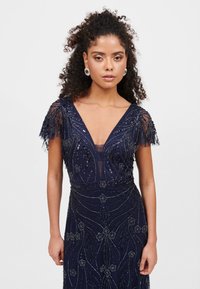 Námořnická modrá šaty s hlubokým výstřihem do V, korálkovými květinovými vzory, průsvitnými rukávy a přiléhavým střihem. Texturovaná tkanina se třpytivými akcenty.