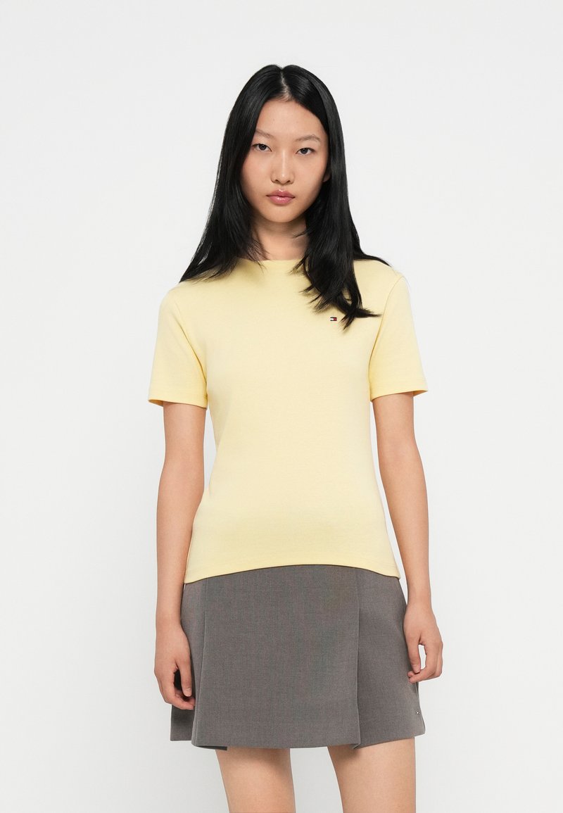 Gul kortermet T-skjorte i mykt stoff med rund hals, brukt sammen med et grått skjørt med et subtilt mønster. Logofokus på skjorten.