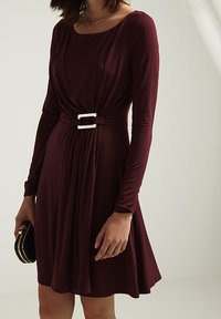 Bordowa sukienka z długim rękawem ma dopasowaną górę, pasek w talii z srebrną klamrą oraz rozkloszowaną spódnicę z fałdami.
