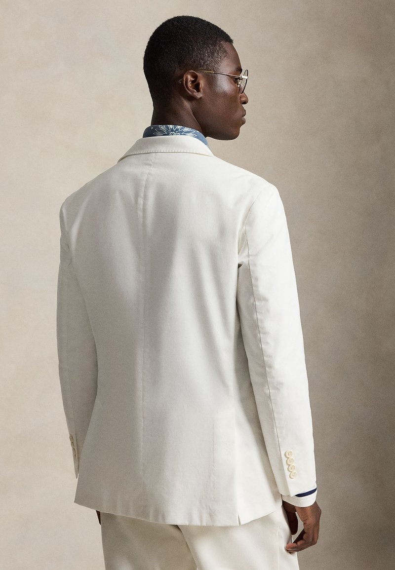 Homme portant une veste de costume blanche et une chemise à motifs, de profil avec un fond beige neutre.