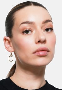 Orecchini a cerchio in oro con un design texture, caratterizzati da un cerchio più piccolo intrecciato. Il soggetto presenta un trucco naturale e una pelle liscia.