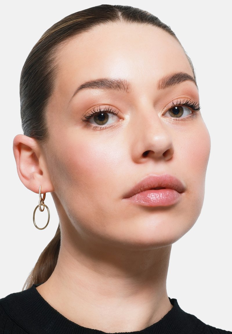 Orecchini a cerchio in oro con un design texture, caratterizzati da un cerchio più piccolo intrecciato. Il soggetto presenta un trucco naturale e una pelle liscia.