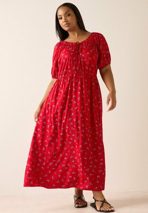 Mujer vestida con un vestido rojo hasta los tobillos con pequeños estampados florales y mangas abullonadas, de pie frente a una pared lisa y clara.