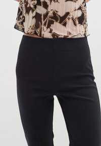 Zwarte fitted broek met elastische tailleband, gecombineerd met een doorschijnende blouse met een bloemenpatroon in beige en bruin.