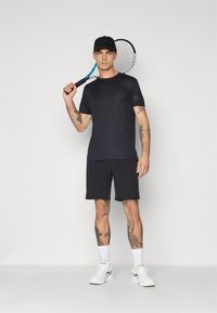 Μαύρη αθλητική μπλούζα και σορτς, συνδυασμένα με λευκά αθλητικά παπούτσια. Το μοντέλο κρατάει ένα ρακέτα τένις και φοράει μια μαύρη καπέλα με λογότυπο.