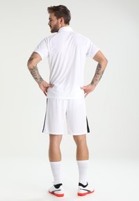 Maglietta sportiva bianca con colletto, abbinata a pantaloni corti bianchi con strisce nere ai lati, calze bianche e scarpe da ginnastica con dettagli rossi.