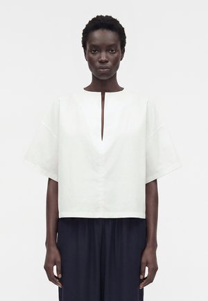 Γυναίκα με κοντά, σγουρά μαλλιά στέκεται με το πρόσωπο προς τα εμπρός, φορώντας έναν άνετο λευκό κοντομάνικο τοπ με άνοιγμα στο ντεκολτέ μπροστά και σκούρο παντελόνι με φαρδιά παντελόνα.