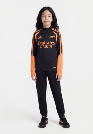 Enfant debout portant une tenue de sport Arsenal noire et orange avec le logo "Emirates Fly Better" et des baskets assorties noires et orange.