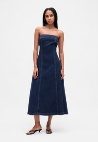 Tamno traper haljina bez naramenica s pripijenim gornjim dijelom i lepršavom suknjom A-kroja. Ima vidljive šavove i rub koji seže do koljena.