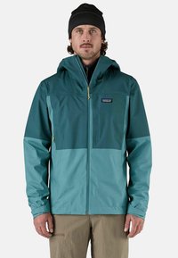 Giacca impermeabile teal con cappuccio, caratterizzata da un design bicolore, chiusura con zip e polsini elasticizzati. Il materiale sembra liscio e leggero.