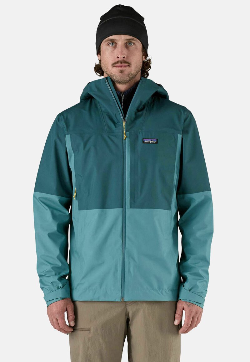 Giacca impermeabile teal con cappuccio, caratterizzata da un design bicolore, chiusura con zip e polsini elasticizzati. Il materiale sembra liscio e leggero.