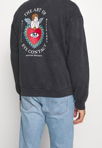 Svart sweatshirt med grafisk design på ryggen, som visar en cherub, ett hjärta och ett öga. Ribbad mudd och nederkant. Kombinerad med ljusblå jeans.