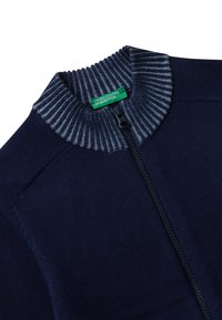 Námořnický svetr na zip s šedým žebrovaným límcem. Obsahuje zelenou značkovou nášivku, hladký povrch a čisté švy.