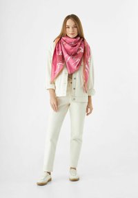 Écharpe rose à motifs en tissu doux, stylée en forme triangulaire, portée par-dessus une veste et un pantalon de couleur claire.