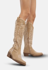 Botas de cowboy beige con intrincadas costuras blancas, diseños decorativos, cremalleras laterales y un bajo tacón de madera. Acabado en cuero suave.