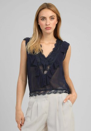 Femme blonde porte un chemisier sans manches en dentelle bleu marine avec des volants et un pantalon blanc plissé taille haute, debout devant un fond gris uni.