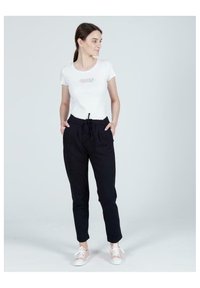 Biała koszulka polo z krótkim rękawem z napisem "CROSS JEANS" w kolorze srebrnym, połączona z czarnymi spodniami dresowymi ze sznurkiem i białymi trampkami.