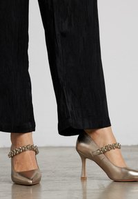 Voeten in metallic puntige hakken met juwelen straps, gecombineerd met zwarte gestructureerde wijde broeken, staande op een grijze vloer.