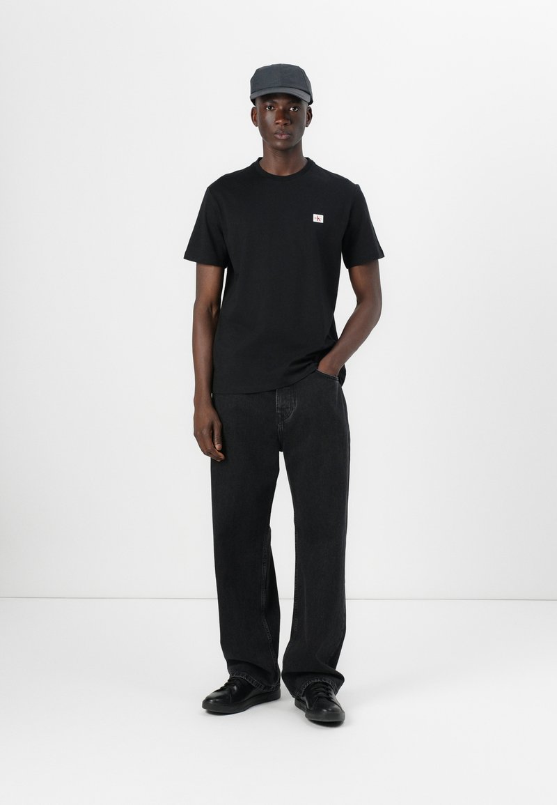 Zwart katoenen T-shirt met een klein logo, gecombineerd met losse zwarte spijkerbroek en zwarte schoenen. Het model draagt een grijze pet.