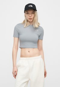 Ξανθιά γυναίκα που φοράει ανοιχτό μπλε κοντομάνικο μπλουζάκι The North Face, λευκή φόρμα και μαύρο καπέλο με το λογότυπο The North Face, στέκεται μπροστά σε λευκό φόντο.