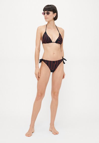 Melnas bikinis komplekts ar trīsstūra formu augšdaļu un saistījuma apakšdaļu, ar drosmīgiem sarkaniem rakstiem. Modelis valkā rozā tonētus saulesbrilles.
