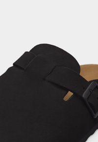 Nahaufnahme einer schwarzen Ledersandale mit einem breiten Riemen und verstellbarer Schnalle auf einem hellbraunen Fußbett.