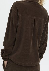 Bruin corduroy overhemd met een achterkantplooi, lange mouwen en opgerolde manchetten. Gelaagd stof met verticale ribbels, minimalistisch ontwerp.
