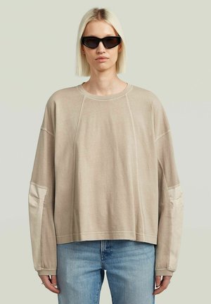 Personne aux cheveux blonds raides portant des lunettes de soleil noires, une chemise beige oversize à manches longues avec des coudières claires et un jean bleu.