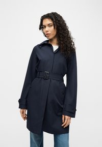 Νεαρή γυναίκα με σγουρά μαλλιά που φοράει ένα μπλε παλτό τραντσέ με ζώνη και μπλε τζιν, όρθια μπροστά σε ένα απλό λευκό φόντο.