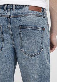 Vue arrière d'une personne portant un jean en denim bleu clair avec des poches arrière visibles et un patch en cuir marron marqué "!SOLID" sur la ceinture.