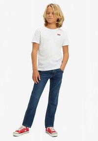 Λευκό κοντομάνικο μπλουζάκι με μικρό κόκκινο λογότυπο, συνδυασμένο με μπλε τζιν και κόκκινα αθλητικά παπούτσια με λευκές λεπτομέρειες. Στάση όρθια, με τα χέρια στις τσέπες.