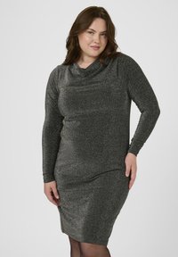 Femme grande taille portant une robe pailletée grise foncée à manches longues et à hauteur des genoux, posant avec une main sur la hanche sur un fond uni.