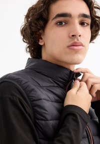 Chaleco negro acolchado con un cuello alto y costuras horizontales, que presenta una cremallera naranja. Lleva puesto una camiseta negra de manga larga.