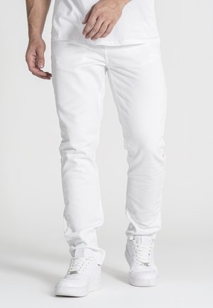 Man wearing white pants, white sneakers, and a white shirt standing against a plain light background.