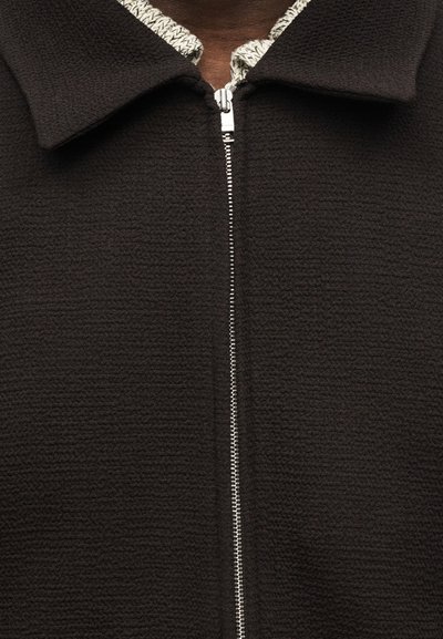 Σκούρο υφασμάτινο σακάκι με ασημένιο φερμουάρ μερικώς κλειστό πάνω από πλεκτό λευκό και μαύρο μοτίβο μπλουζάκι που φαίνεται στον γιακά.