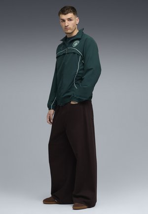 Grüne Windjacke mit weißen Paspeln und einem Logo. Schwarze weite Hosen. Braune Schuhe sind sichtbar. Einfaches Design und glatte Oberfläche.