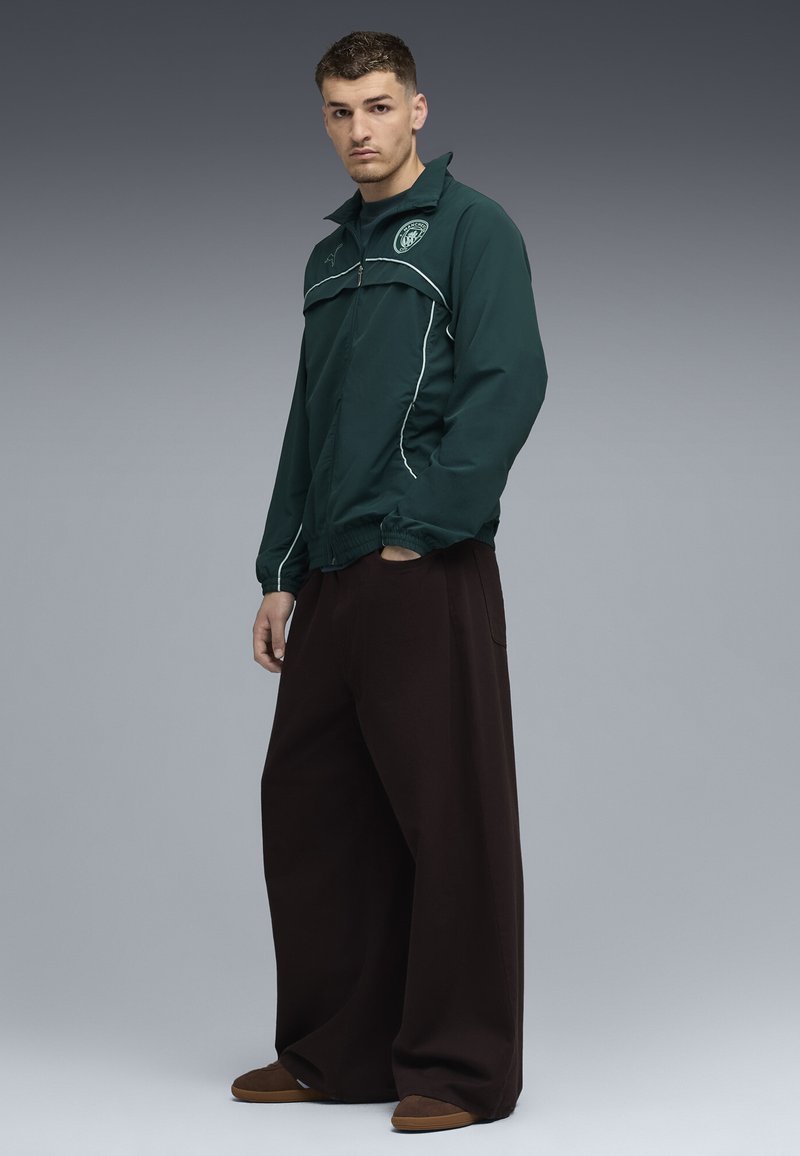 Grüne Windjacke mit weißen Paspeln und einem Logo. Schwarze weite Hosen. Braune Schuhe sind sichtbar. Einfaches Design und glatte Oberfläche.