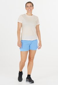 Beige svedtransporterende skjorte med korte ærmer, parret med lyseblå sportsbukser. Modellen har sorte løbesko på. Simpelt design, ingen mønstre.