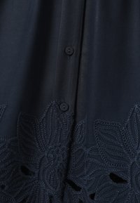 Tissu sombre avec deux petits boutons et un motif de dentelle florale brodé le long du bord, représentant des formes de feuilles et de pétales.