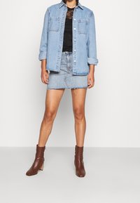 Camicia di jeans azzurro chiaro con due tasche sul petto, abbinata a un top di pizzo scuro e a una gonna di jeans corta e sfrangiata. Stivaletti alla caviglia marroni.